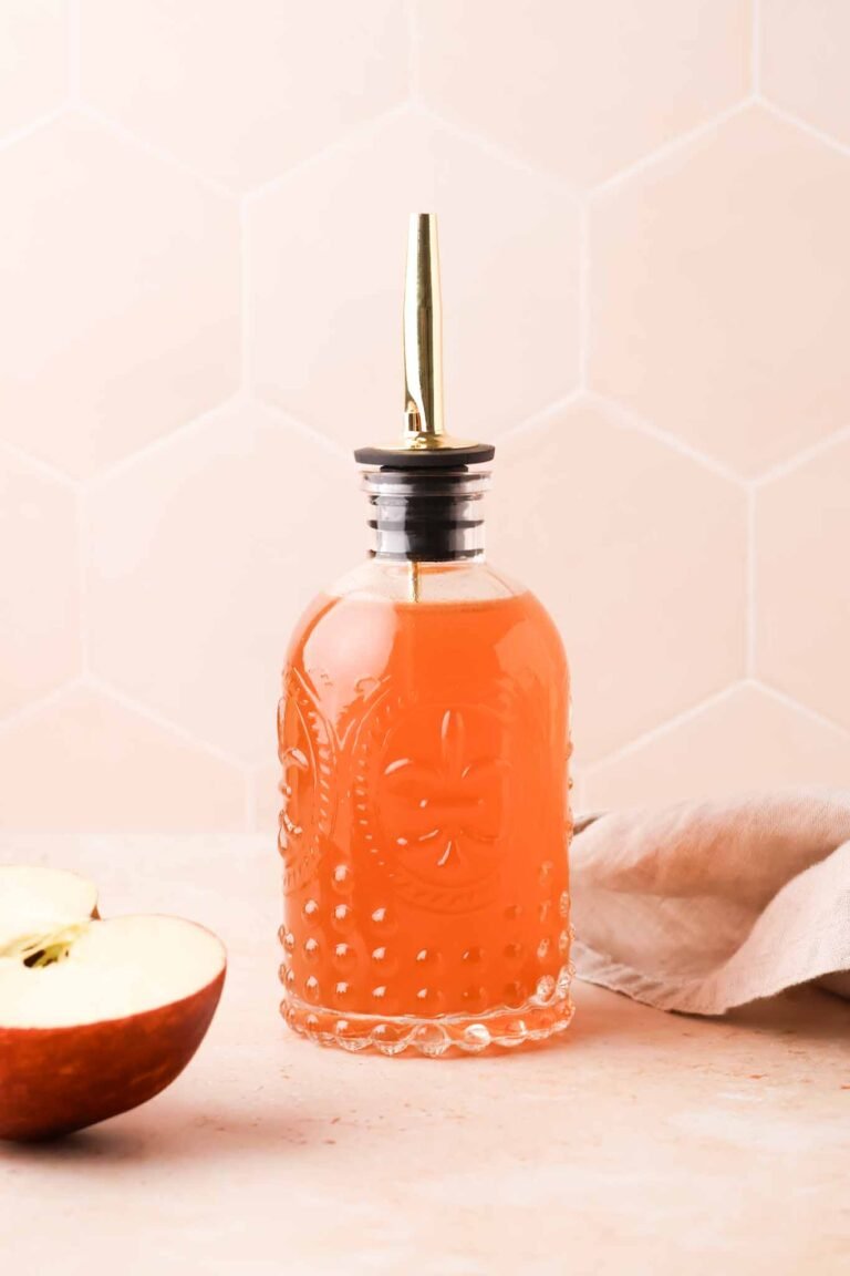 apple cider syrup in a glass bottle sitting on a table, surrounded by sliced apples