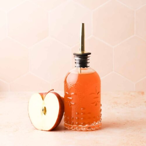 apple cider syrup in a clear glass bottle on a table, with apple halve nearby
